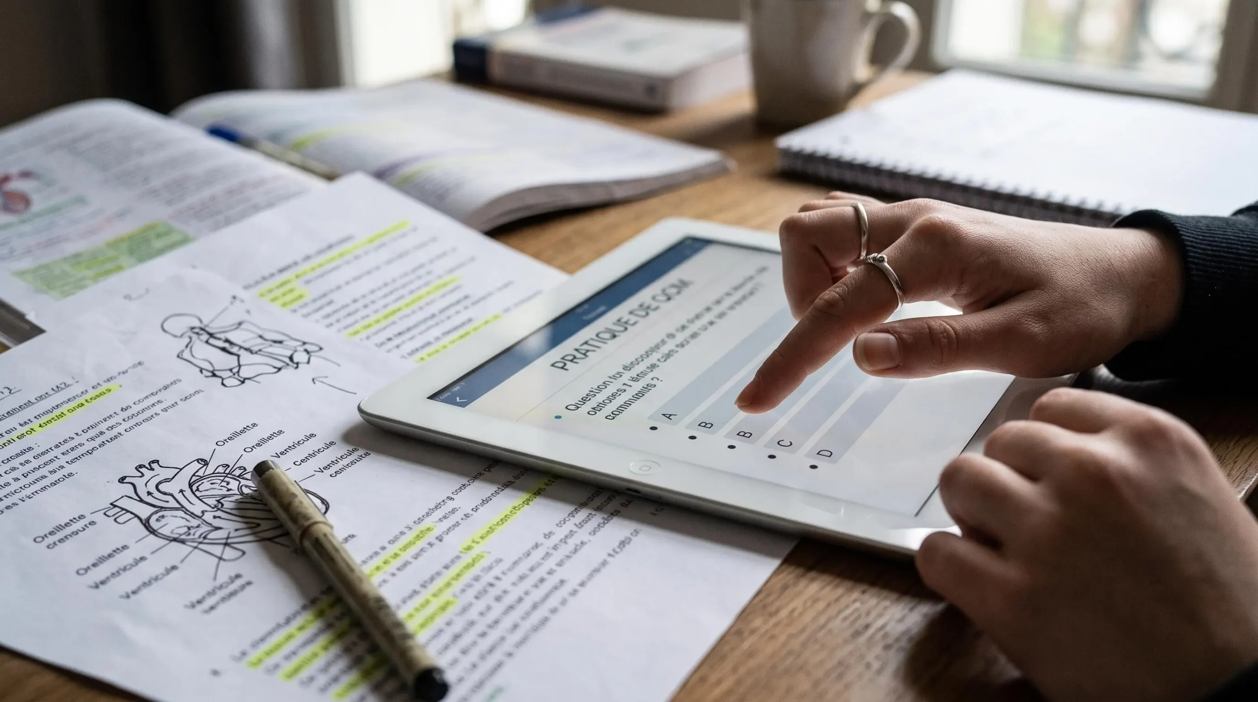 Mains d'un étudiant LSPS sélectionnant une réponse sur un QCM de tablette numérique avec cours médical à proximité